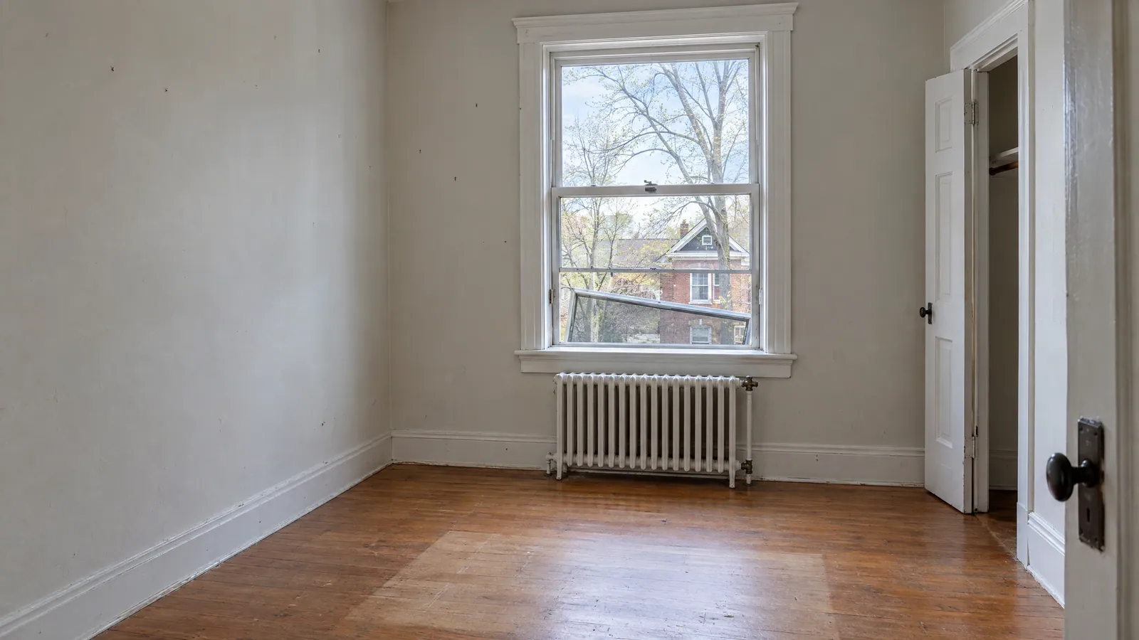 Empty bedroom in an older rental house with scuffed walls, a radiator and a double-hung window whose window screen hangs bent out of its track