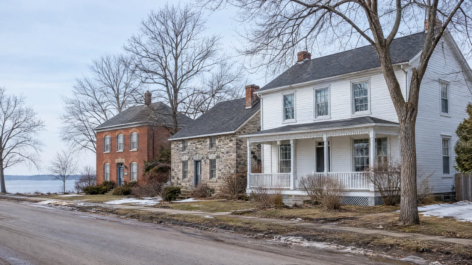 Village street in late winter with a red brick house, a fieldstone house and a white clapboard house with a porch, all with multi-pane wood sash windows, the lake beyond